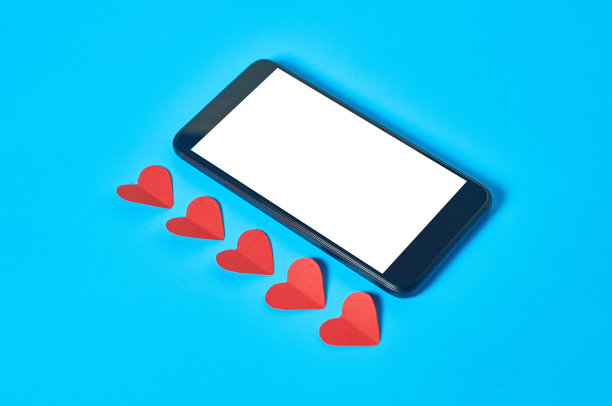 黑色智能手机与孤立的白色屏幕文本，图片，照片和其他图形附近的红色纸心在蓝色的桌子。情人节和爱情概念。空间的文本图片下载