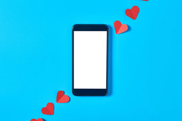 黑色智能手机与孤立的白色屏幕文本，图片，照片和其他图形附近的红色纸心在蓝色的桌子。情人节和爱情概念。空间的文本。俯视图图片下载