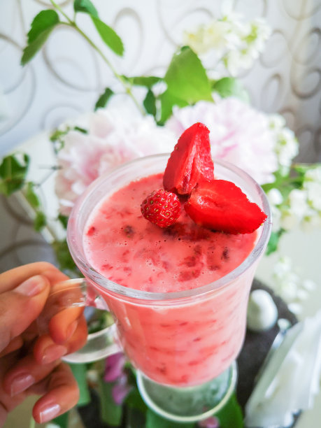 一个女孩手里拿着草莓奶昔。女人喝着粉红色的浆果鸡尾酒，鸡尾酒的背景是花，垂直的照片。图片下载