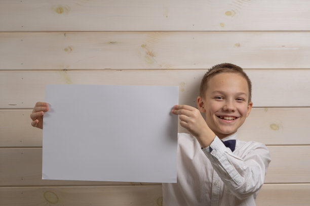 一个10岁的男孩兴高采烈地拿着一张白纸，背景是一堵木墙。文本。模型。图片下载