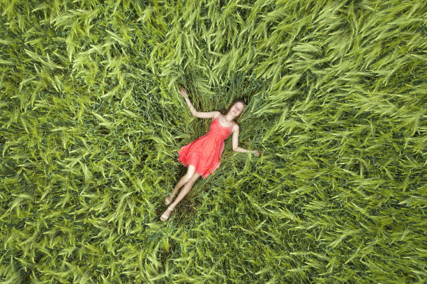 从上面看，一个年轻迷人的苗条女人穿着红色的衣服，闭着眼睛躺在绿色的麦田里。图片下载