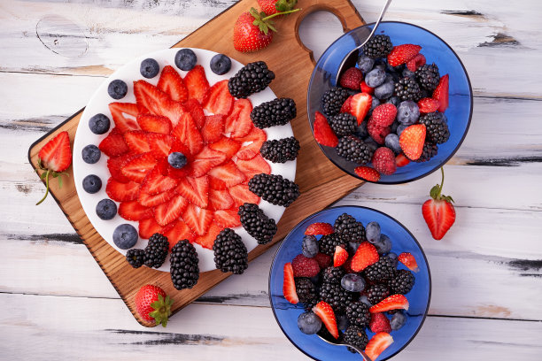 蓝莓、覆盆子、黑莓和草莓片在一个白色盘子里围成一个圈，然后混合在蓝色的碗里，放在砧板上，背景是浅色的木头。素食菜单，健康食物。从上面的观点图片下载