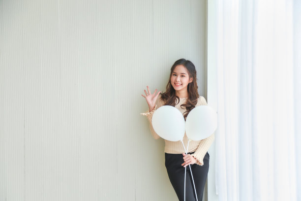 肖像图片美丽的年轻亚洲女人在米色休闲衬衫拿白色气球，使一只手说嗨，看着相机和米色墙微笑。图片下载