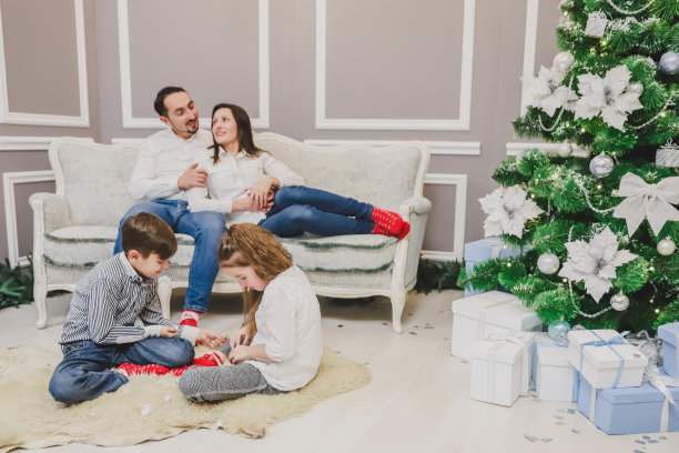 照片中的爸爸、妈妈和女儿在家里的圣诞树旁，都面带微笑。孩子们在最前线玩耍。图片下载