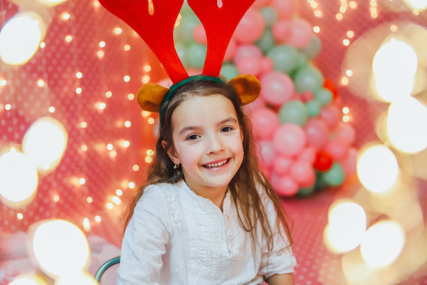 散景的灯光从圣诞花环上走在最前面，可爱的小女孩拿着驯鹿角，看着镜头，在晚上微笑着在家。图片下载