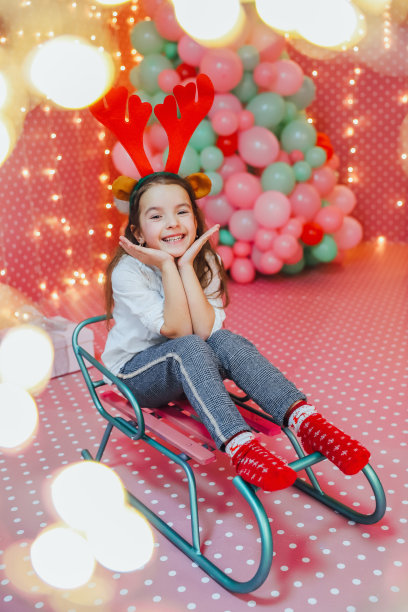 可爱的小女孩坐在粉红色的房间里的雪橇上，气球和散竹花环在最前面。图片下载