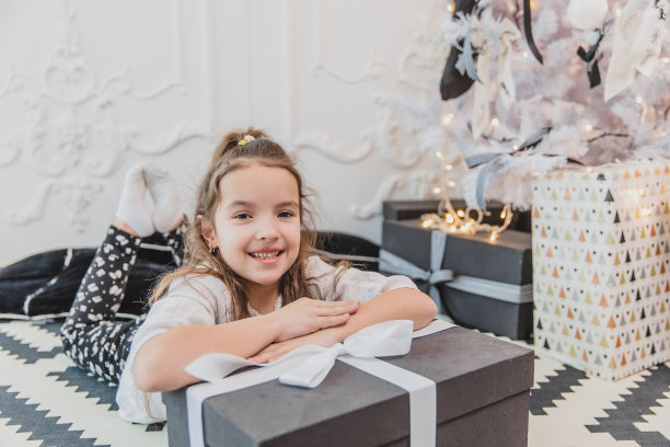 睡眼惺松但快乐的孩子穿着圣诞睡衣躺在圣诞树下的地板上，手一直放在大礼物上。图片下载