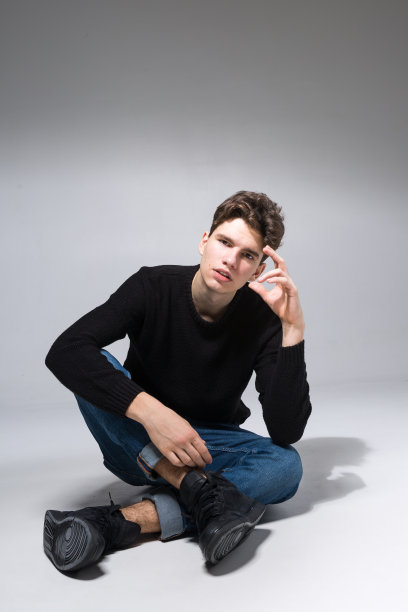 一个穿着牛仔裤和黑色运动衫坐在地板上摆造型的时尚男士。男学生肖像全白色背景。摄影:英俊迷人的欧洲男子坐在地板上图片下载