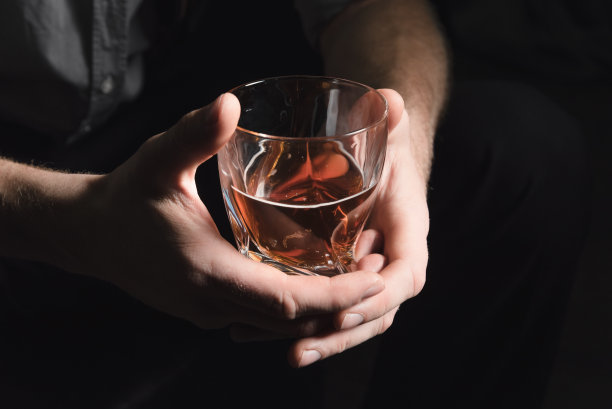 一个年轻人的手的特写，谁倒酒精从酒瓶到一个玻璃杯，在一个黑暗孤立的背景图片下载