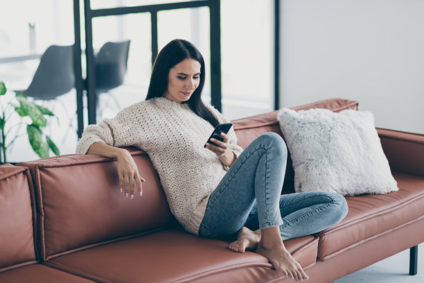 简介侧面的照片集中的女孩用她的智能手机，与朋友在线聊天，写短信，坐在棕色的皮革沙发在家里，室内，穿针织毛衣牛仔牛仔裤图片下载