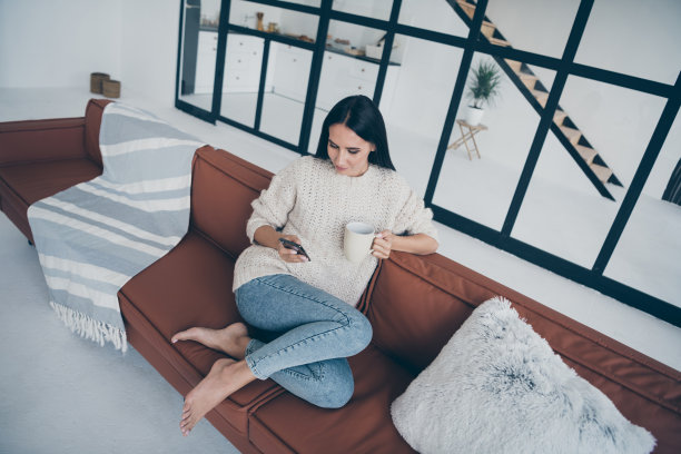 上面高上面的角度，集中的女孩的照片，坐在棕色的皮革沙发上，用手机看新闻，发帖评论，拿着杯，拿着拿铁，穿针织套衫，牛仔，牛仔裤在家里度假在室内图片下载