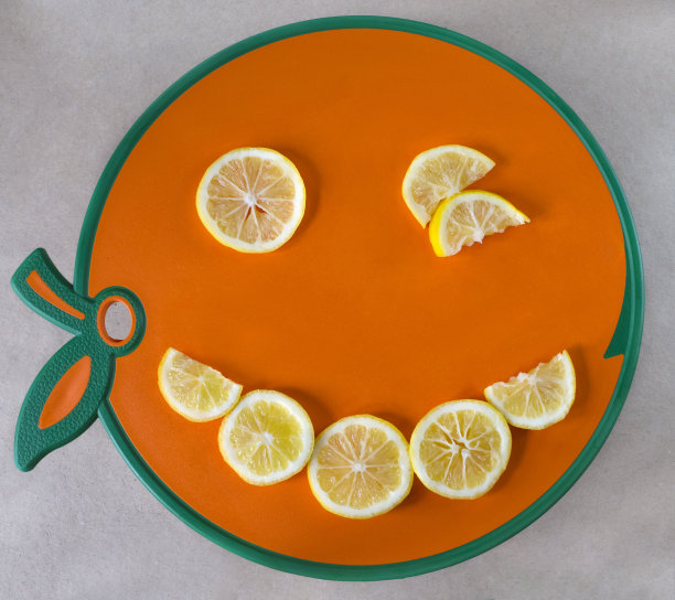 切片的黄色柠檬水果以笑脸的形式摆放在橘子砧板上。孤立在灰色背景。图片下载