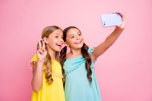 照片中的两个金发褐发小女士模特同学拿着电话做v标志夏季自拍穿亮蓝色的黄色衬衫连衣裙孤立的粉色背景图片下载
