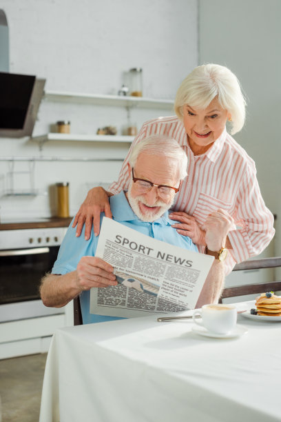微笑的资深妇女拥抱激动的丈夫与报纸在厨房早餐图片下载