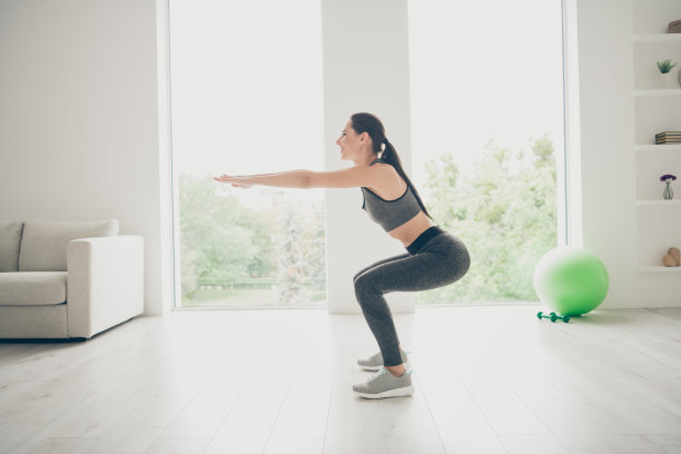 快乐的女孩运动员做运动，坐，蹲，伸手，想成为强壮的幸福的女人穿着灰色的裤子在房子里像健身房一样图片下载