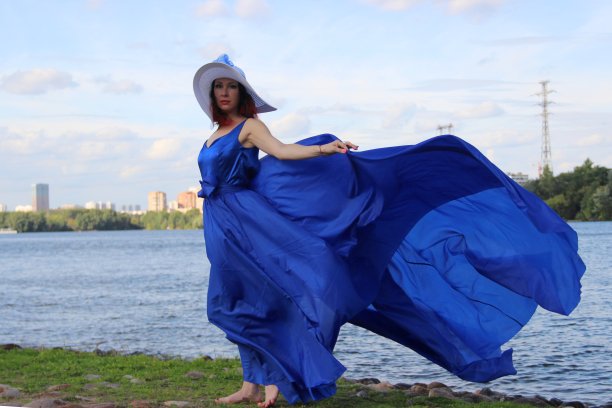 一头红卷发的女人，穿着随风飘动的蓝色长裙，戴着一顶宽边白帽子。沿着河边散步图片下载