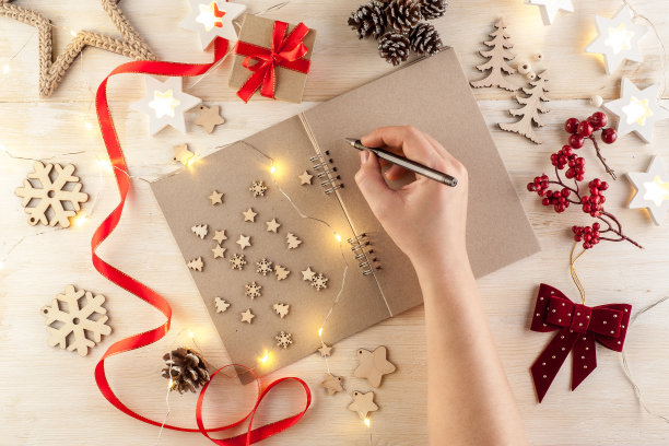 在一张木桌上打开一个用黑色纸包着的笔记本，桌子周围是手工制作的圣诞装饰品和一个花环。图片下载