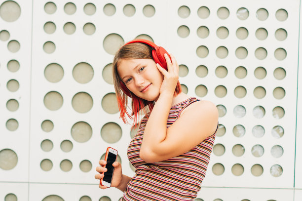 户外肖像快乐的年轻女孩与时尚的红色染短发，孩子听音乐耳机，拿着手机图片下载