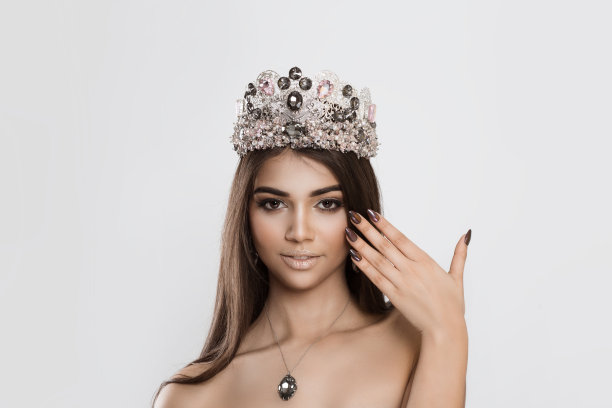 美丽和快乐。美丽加冕的女王女孩女演员小姐新娘是手近脸微笑显示美甲看着相机白墙。全妆钻石银黑粉水晶皇冠图片下载