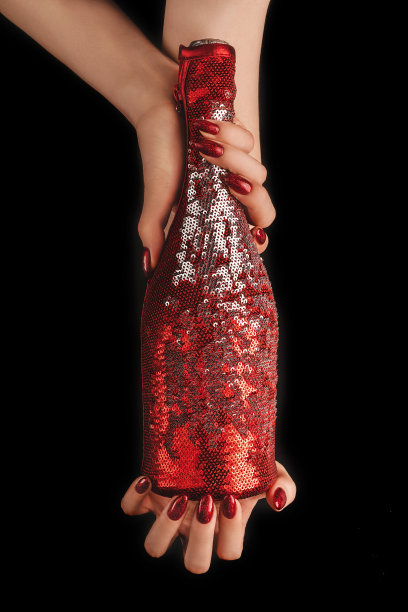 美丽的女性手与时尚的红色美甲持有香槟瓶在封面上闪耀着充满活力的红色亮片在孤立的黑色背景。模型为您的设计。副本的空间。平的。图片下载