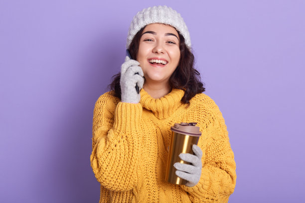肖像欢快的魅力微笑卷发黑发在电话，有积极的面部表情，穿着黄色毛衣，灰色手套和帽子，拿着热杯在手里。图片下载