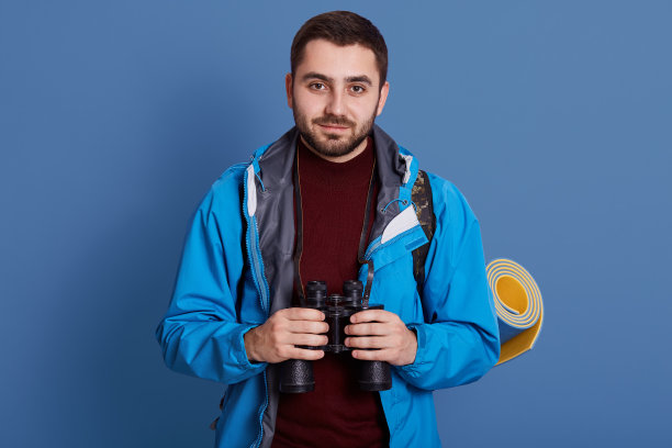 这张照片上，一个兴高采烈的人，脸上有一副愉快的表情，双手拿着望远镜，直视着摄像机，背带着睡袋，穿着露营服。旅行的概念。图片下载