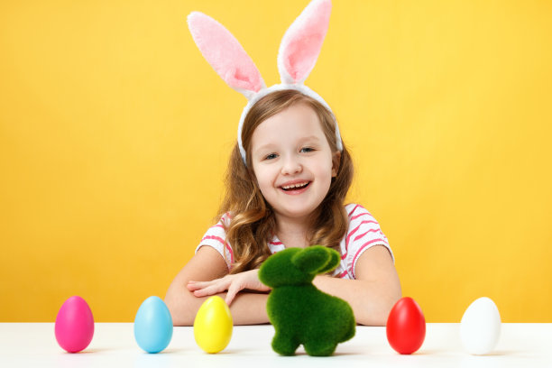 复活节的概念。快乐的孩子兔耳朵黄色背景。小女孩坐在桌前，桌上放着彩绘的鸡蛋和一只兔子图片下载