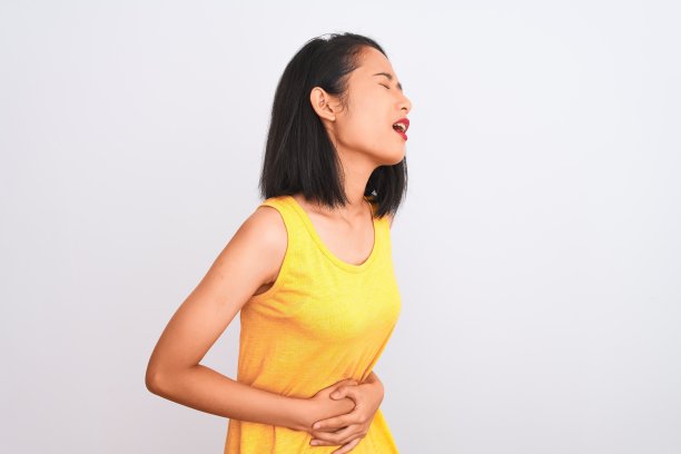 年轻的中国女子穿着黄色休闲t恤站在孤立的白色背景与手对胃，因为消化不良，痛苦的疾病感觉不舒服。疼痛的概念。图片下载