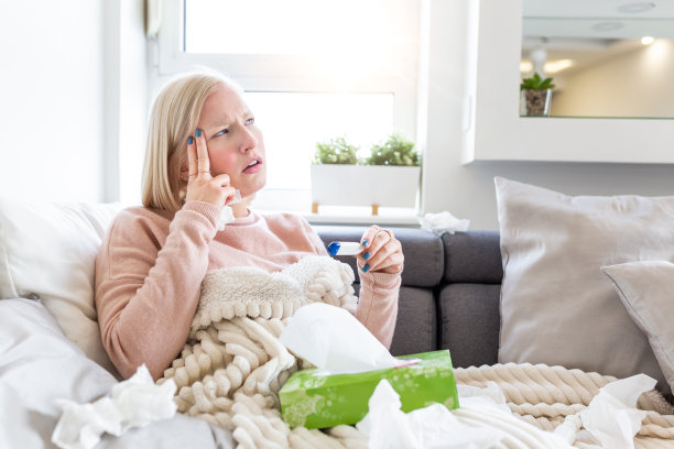 疾病、季节性病毒问题概念。一个得了流感的女人躺在沙发上看温度计上的温度。病人躺在床上发高烧。感冒和偏头痛。素材图片