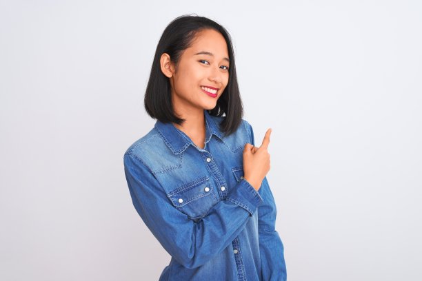 年轻美丽的中国女人穿着牛仔衬衫站在孤立的白色背景愉快地微笑着指着与手和手指向上的一边与快乐和自然的表情在脸上图片下载