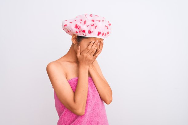 年轻的中国妇女穿着浴巾和帽子在孤立的白色背景与悲伤的表情掩盖脸与手哭泣。抑郁症的概念。图片下载