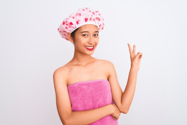 年轻的中国女子穿着浴巾和帽子沐浴在孤立的白色背景上，微笑着，快乐的脸在镜头前眨着眼睛做胜利标志。二号人物。图片下载