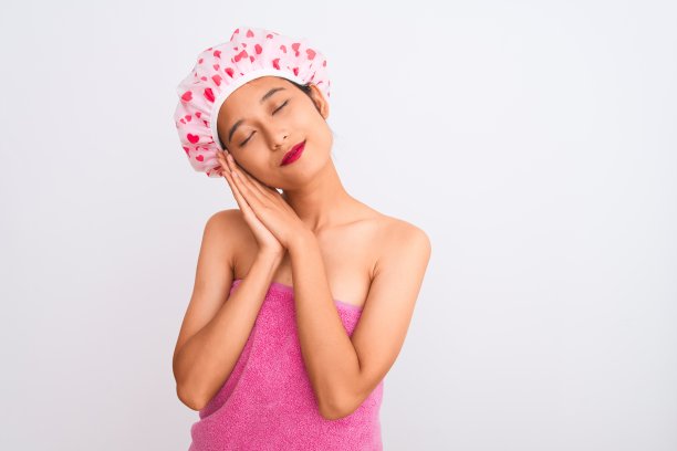 年轻的中国女子穿着浴巾和帽子，在孤立的白色背景下睡觉累了，做着梦，闭着眼睛微笑着双手摆姿势。图片下载