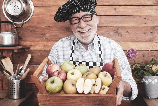 正面的一个有吸引力的和愉快的老年人拿着一个装满各种质量和颜色的苹果篮子。回收木材的乡村背景。一个人只有图片下载