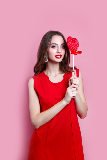 美丽的黑发女孩在红色裙子拿着一个红色的心形棒棒糖在粉红色的背景。情人节图片下载