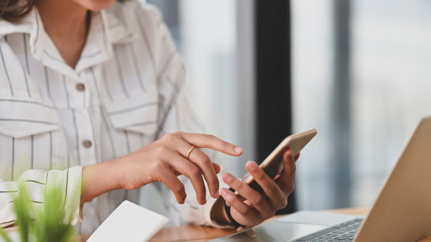 剪短的照片，年轻富有创造力的女人在条纹衬衫拿着和使用她的智能手机在现代的办公桌。图片下载