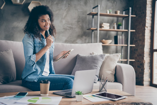漂亮的深色皮肤卷曲的商务女士的照片浏览笔记本桌子，拿着计划表写一周的待办事项，坐在舒适的沙发上，穿休闲牛仔装在室内图片下载