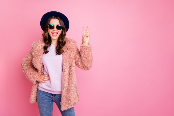 有趣的女士在国外说你好的照片显示v符号外国男人朋友穿着时尚的蓬松的夹克太阳眼镜复古的蓝色帽子孤立的粉色背景图片下载