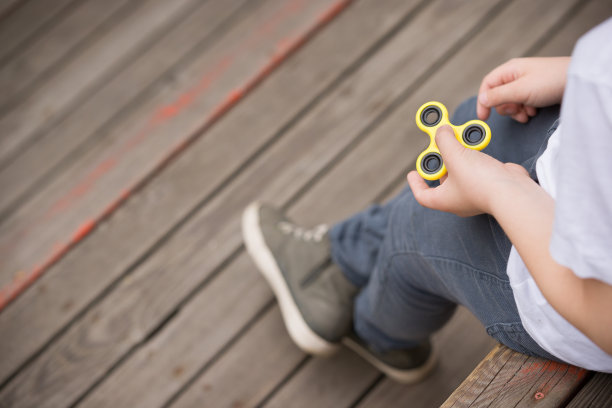小男孩坐在木制操场上玩指尖陀螺——2017年夏季的流行趋势。黄色旋转手，坐立不安的手玩具在孩子的手上旋转素材图片