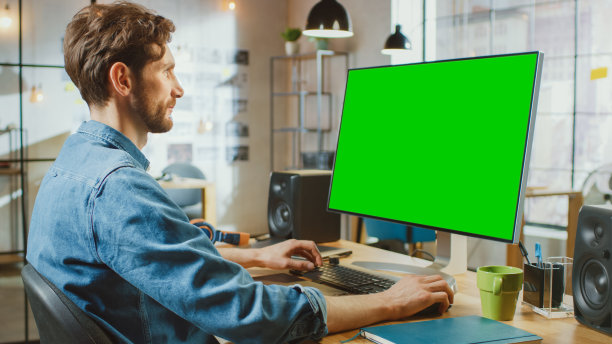 男创意设计师与胡子和牛仔裤衬衫工作在他的个人电脑与大绿色屏幕模拟显示。他在很酷的办公室阁楼工作。图片下载