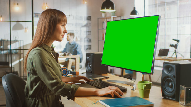美丽的黑发女性视频编辑工作在她的个人电脑与大绿色屏幕模拟显示。她在一个很酷的阁楼办公室工作。其他男性创意同事坐在后台。图片下载