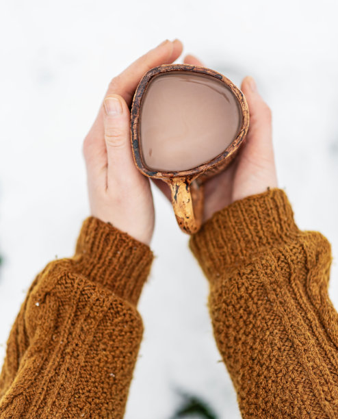穿着针织橙棕色毛衣的女人手捧可可马克杯，背景是雪景图片下载
