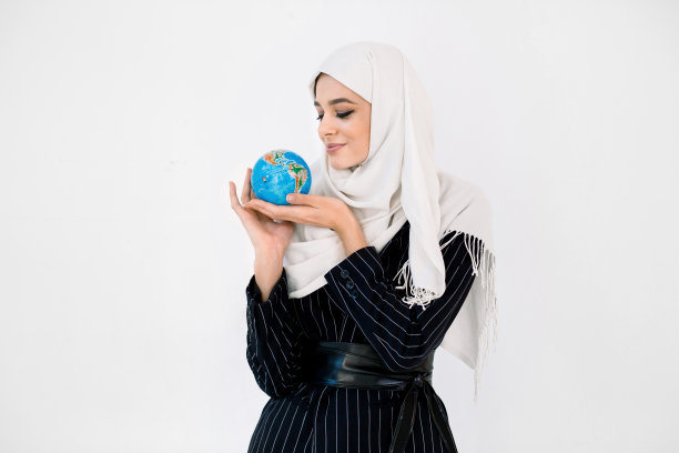 美丽的年轻穆斯林阿拉伯女孩戴着头巾和黑色夹克摆出孤立的白色背景，在棕榈地球世界地球和看地球。地球日。副本的空间。图片下载