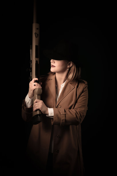 一个穿着棕色斗篷、戴着黑色帽子、手里拿着气枪的女人注视着视线。黑帮风格的照片拍摄。模糊的背景，模糊的焦点，噪音图片下载