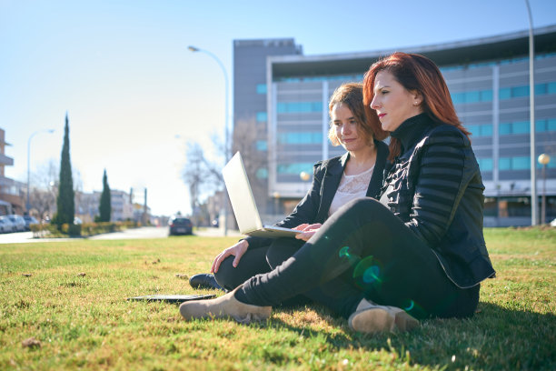 一幅中年妇女的肖像，她的年轻学徒坐在办公楼外的草坪上用笔记本电脑工作。图片下载