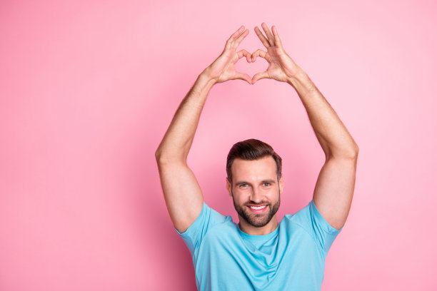 开朗积极的有吸引力的男人的照片，露出牙齿向你展示心形标志与手孤立的时尚柔和的颜色背景图片下载