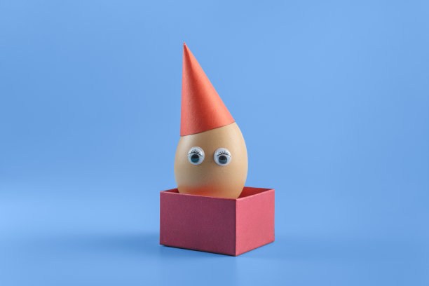 一个棕色的鸡蛋，眼睛戴着红色的节日帽，放在一个方形的红色盒子里，背景是蓝色的。最小复活节概念装饰。图片下载