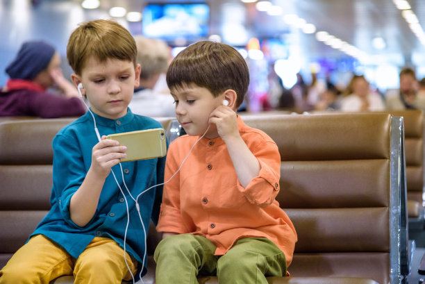 两个小男孩坐在机场的候机大厅里，一边带着行李，一边心满意足地玩着平板电脑或手机。孩子们笑着戴着耳机。带孩子旅行的概念图片下载