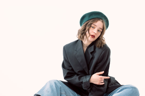戴绿色贝雷帽和黑色西装的时尚卷发女人坐在地上，在白色背景前看着镜头图片下载