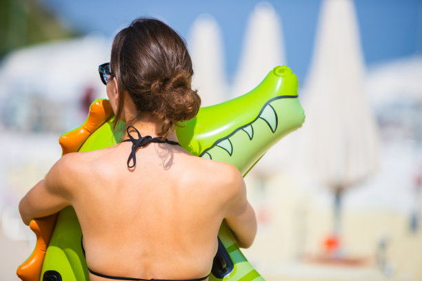 后视图年轻的黑发女人拥抱一个充气鳄鱼或恐龙在海滩上，穿着黑色泳衣图片下载